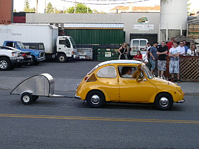 [A small two-door yellow car with a rounded hatchback pulling a tiny silver trailer with a door on its side.]